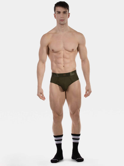A muscular male model stands against a light gray studio background wearing low-rise briefs in olive green, along with black crew socks featuring white stripes.
