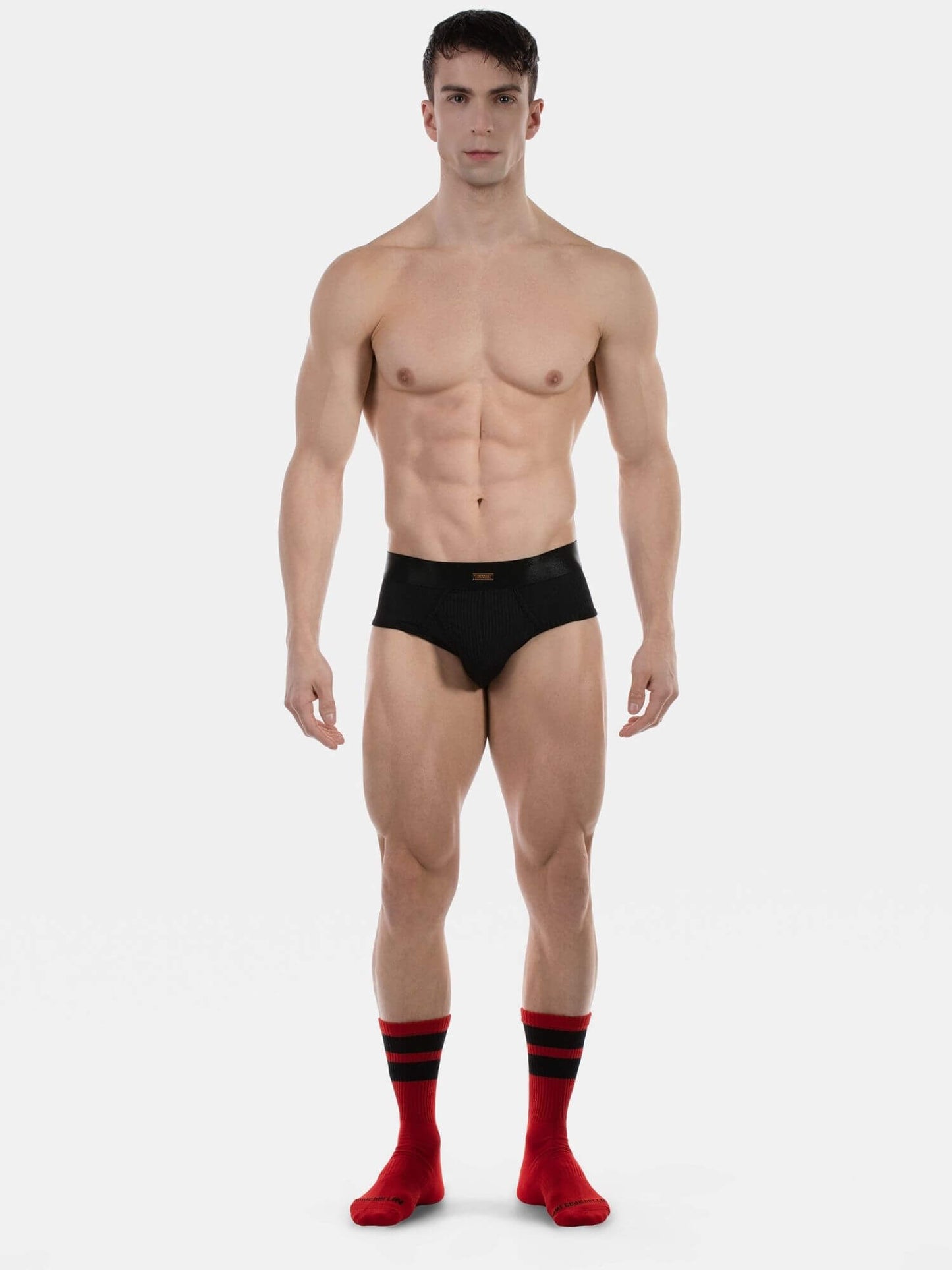 A muscular male model stands against a light gray studio background wearing black briefs and red crew socks with black stripes.