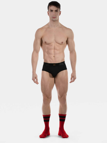 A muscular male model stands against a light gray studio background wearing black briefs and red crew socks with black stripes.