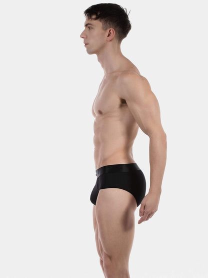 A man in side profile wearing black briefs, photographed against a clean light gray studio background.