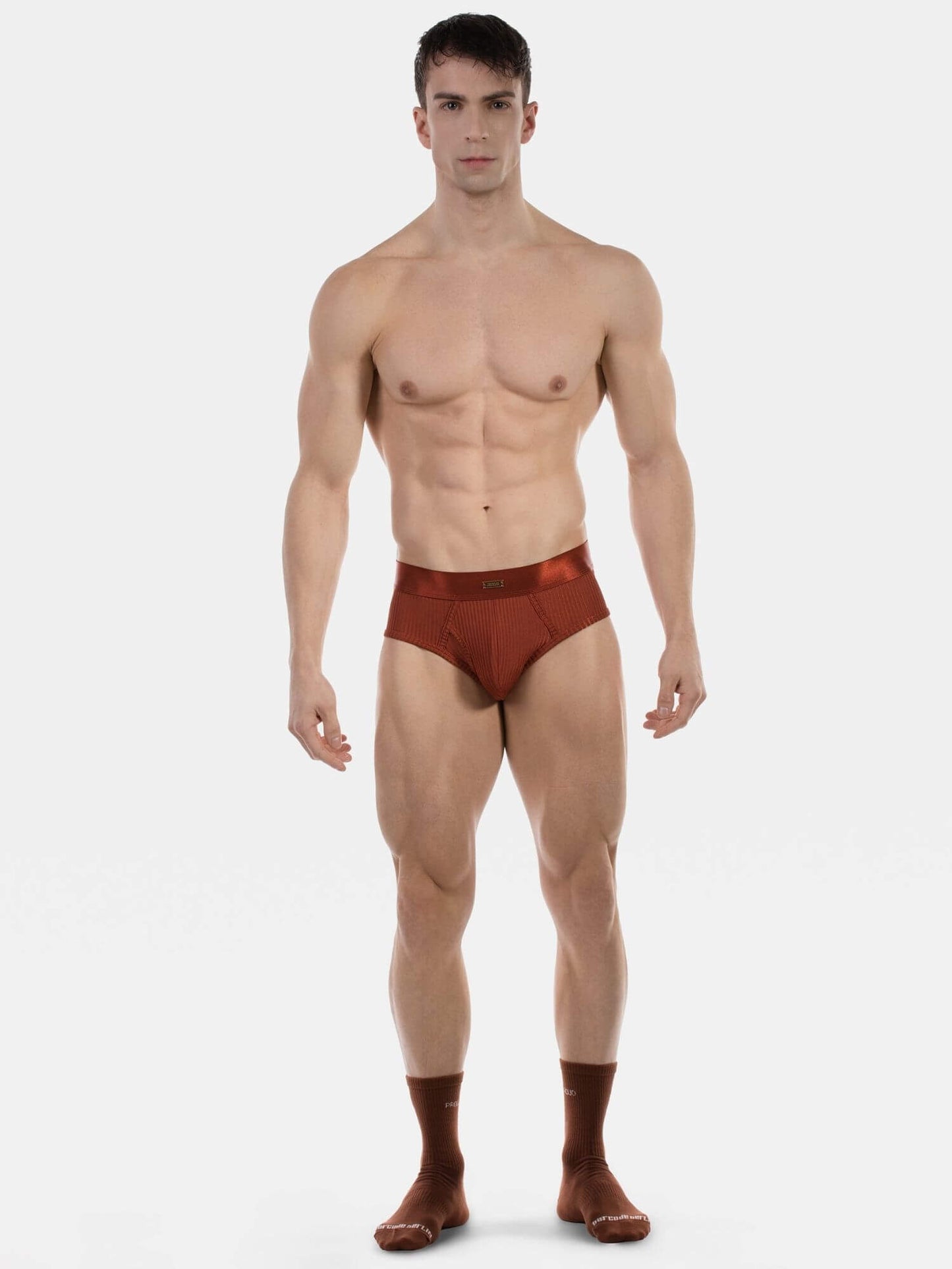 A muscular male model stands against a light gray studio background wearing a pair of low-rise, semi-sheer brown briefs and matching brown socks.