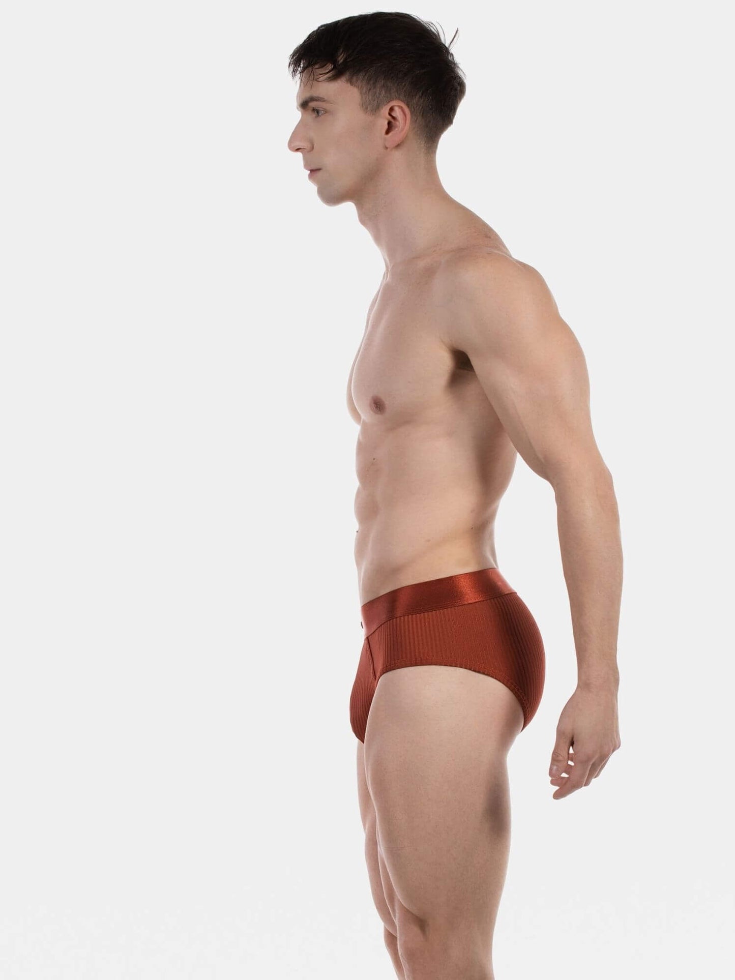 A man in profile wearing low-rise, ribbed copper-brown briefs against a plain light gray studio background.
