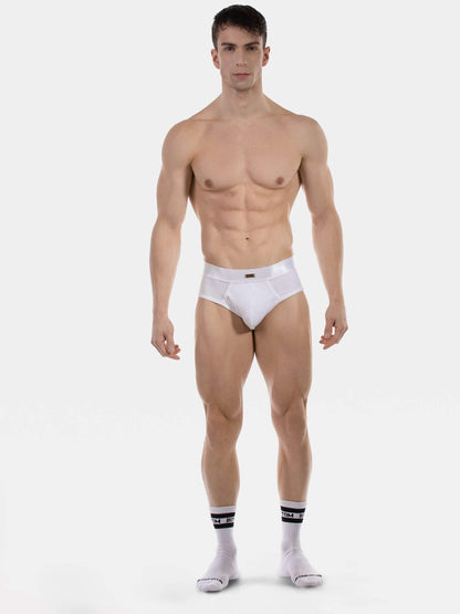 A muscular male model stands against a light gray studio background wearing white briefs and white socks with black stripes.