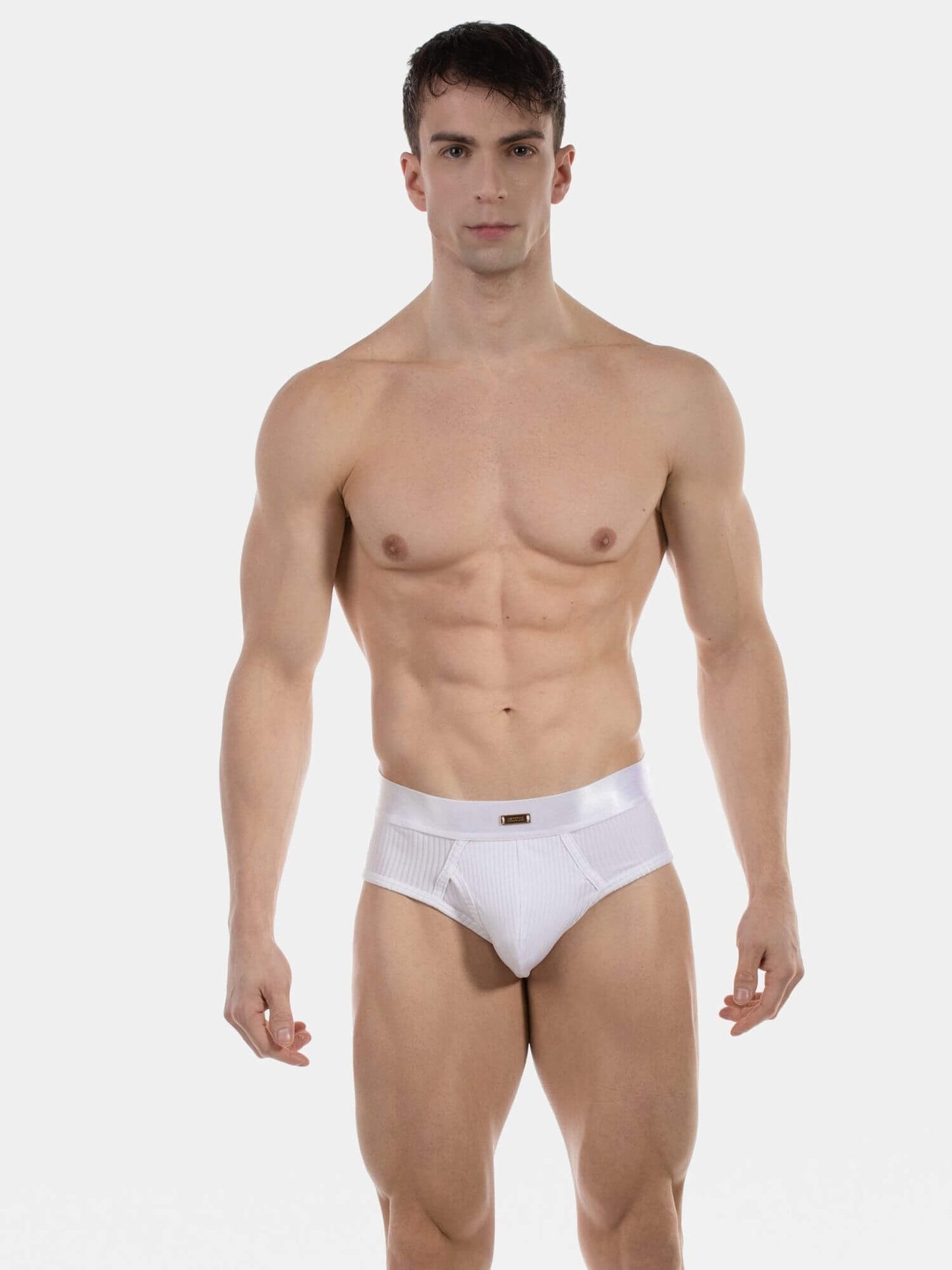 A muscular male model wearing white briefs, standing against a plain light gray studio background.