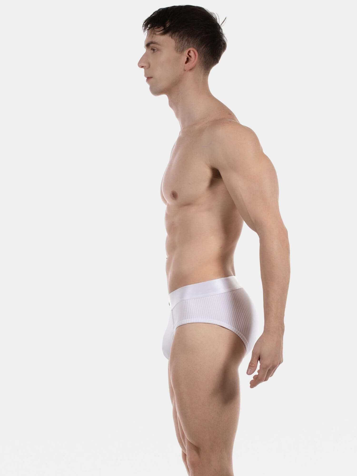 A man shown in side profile wearing white mesh briefs against a plain light gray studio background.