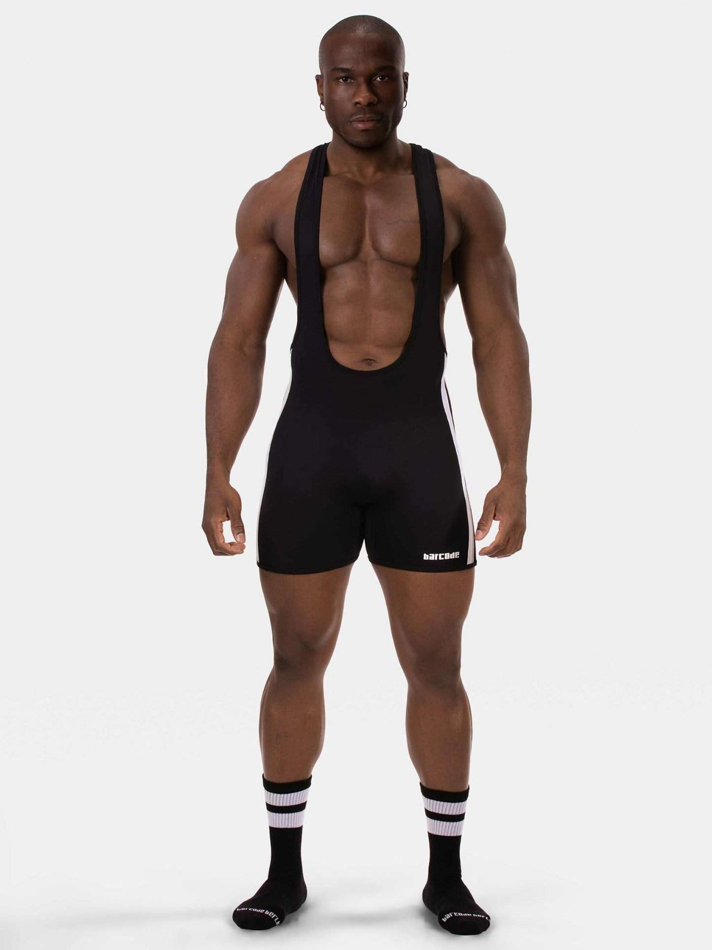 A muscular man modeling a black athletic bib shorts set with suspenders, featuring a small white logo on the left leg, paired with black and white striped socks, against a light gray studio background.