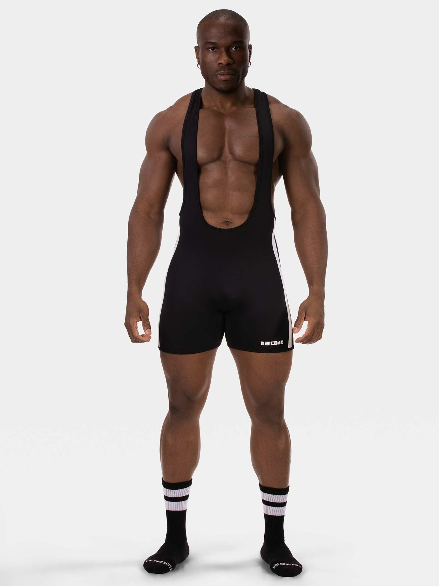 A muscular man modeling a black athletic bib shorts set with suspenders, featuring a small white logo on the left leg, paired with black and white striped socks, against a light gray studio background.
