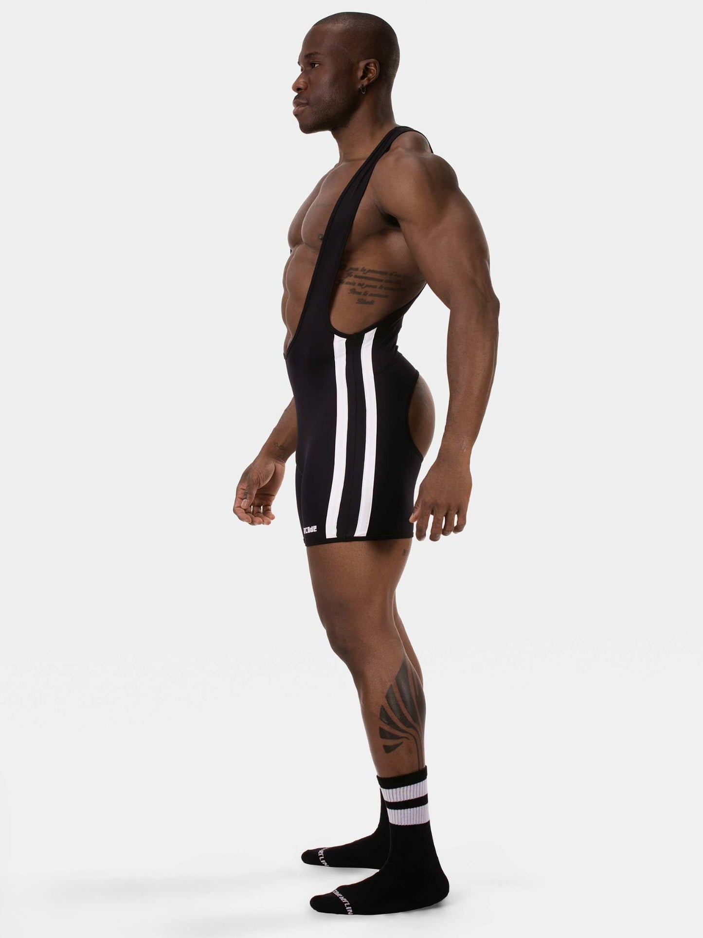 A muscular man in profile wearing a black athletic singlet with three white vertical stripes down the side, paired with black crew socks, against a plain light gray studio background.