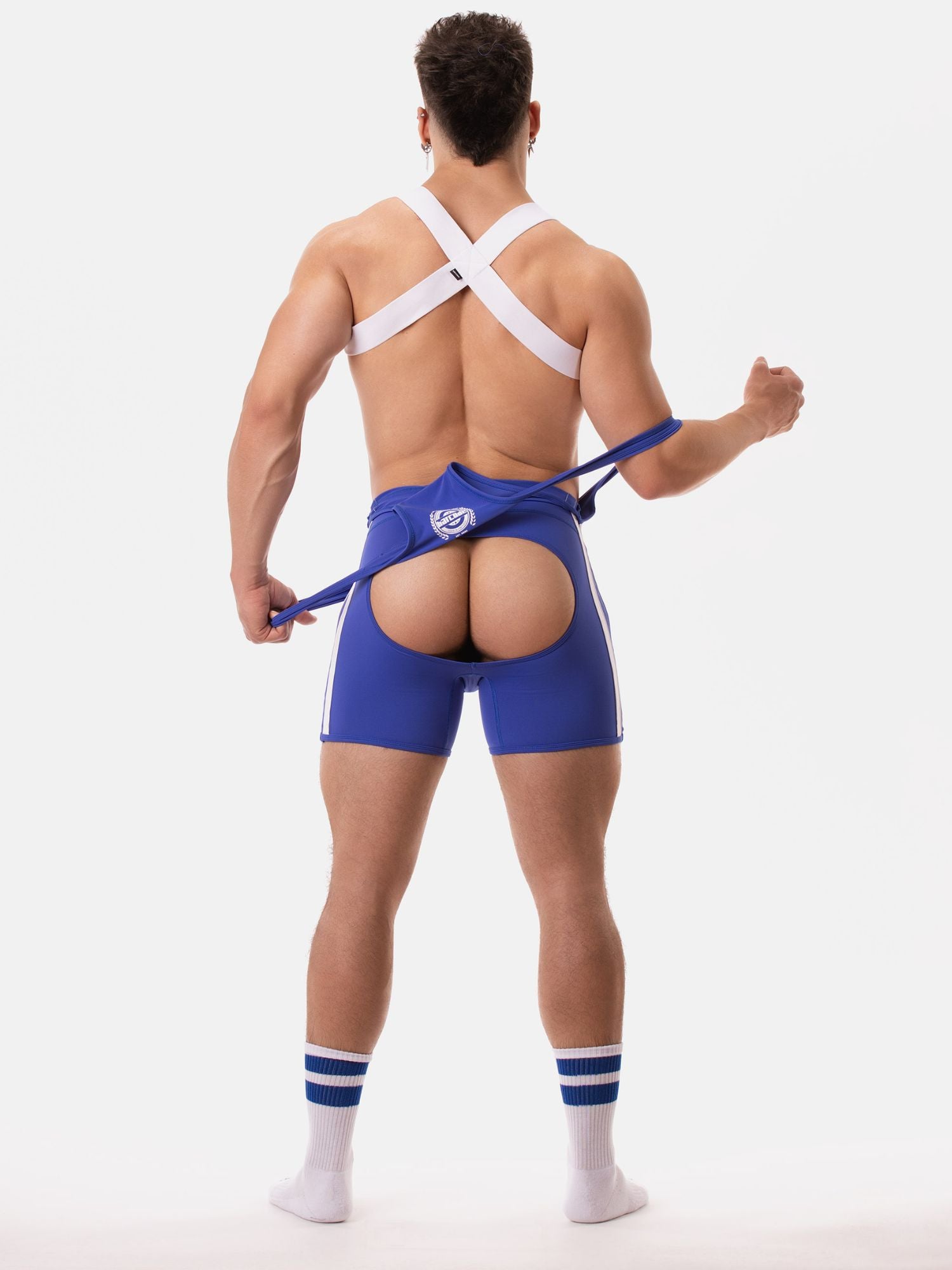 A man shown from behind wearing blue athletic shorts with a large rear cutout, white strappy back support harness, and white socks with blue stripes, against a plain light gray studio background.