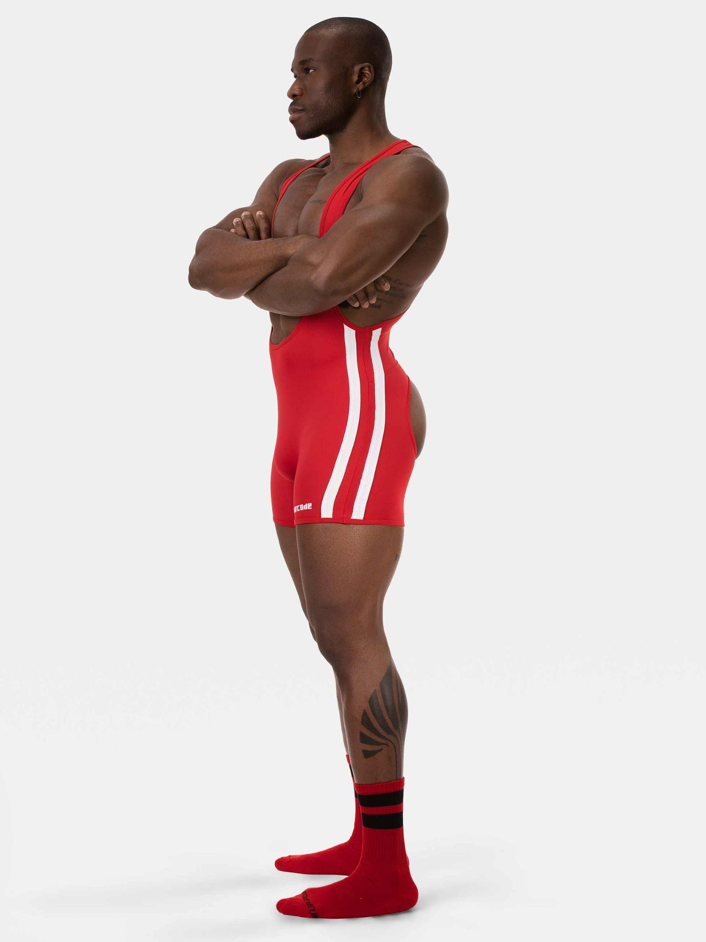 A muscular man stands in profile wearing a red wrestling singlet with white side stripes, red socks, and a black-and-white Adidas logo near the hem, photographed against a light gray studio background.