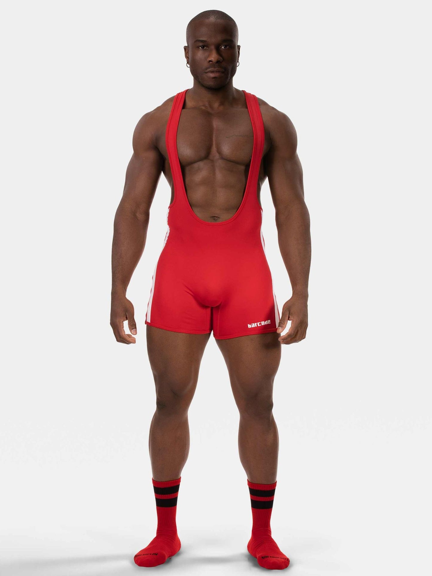 A muscular male model wearing a bright red athletic singlet with shoulder straps and matching red socks with black stripes, standing against a light gray studio background.