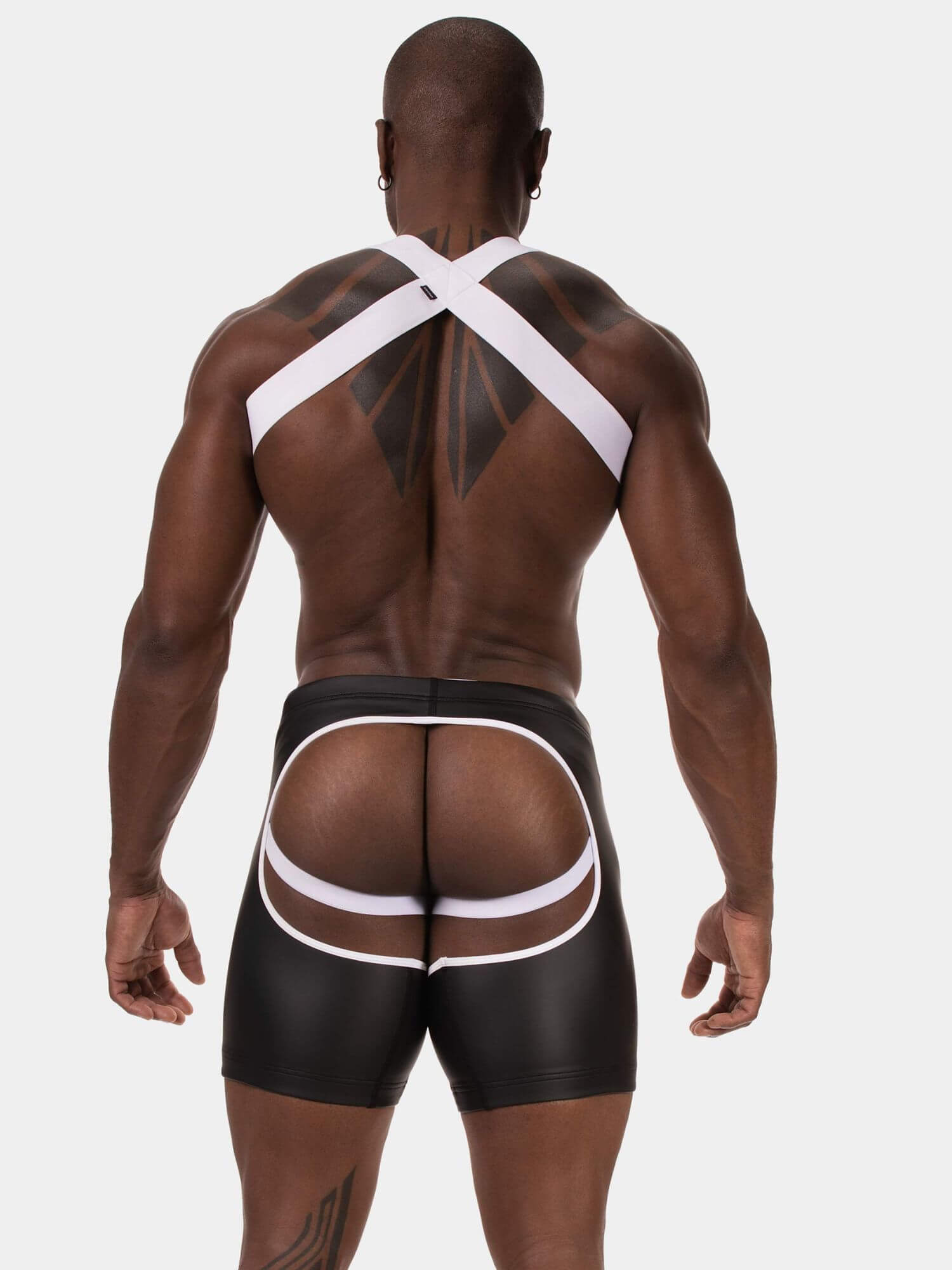 Rear view of a muscular man wearing black briefs with white cutout straps and a white crisscross back strap, against a light gray background.