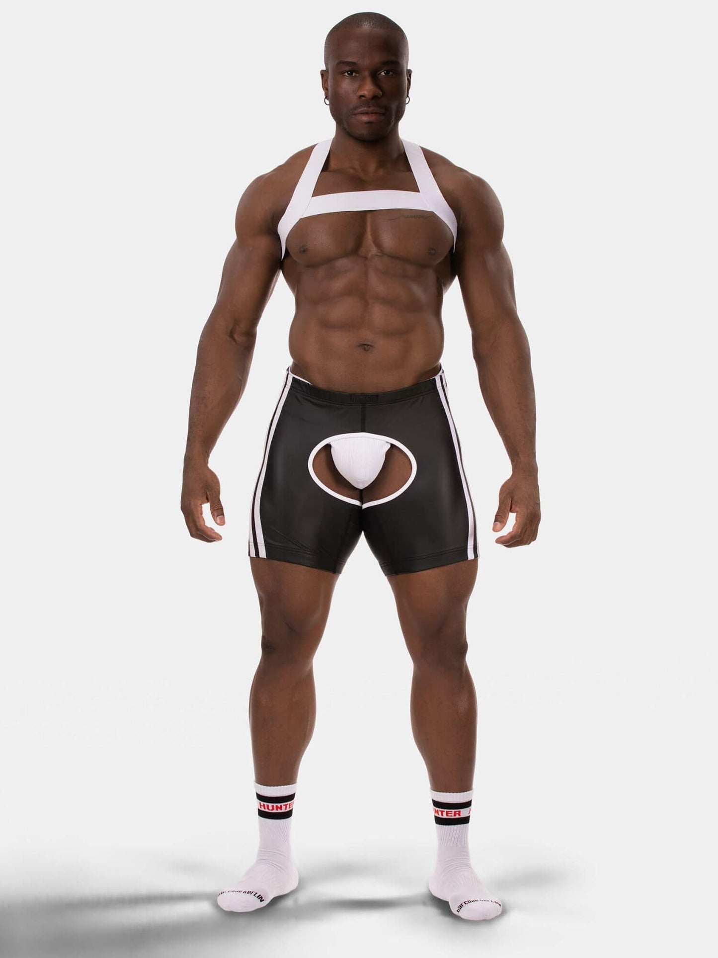 A muscular male model stands against a light gray studio background wearing a white halter-style harness top and black athletic shorts with white side stripes and a white circular front pouch. He is also wearing white socks.