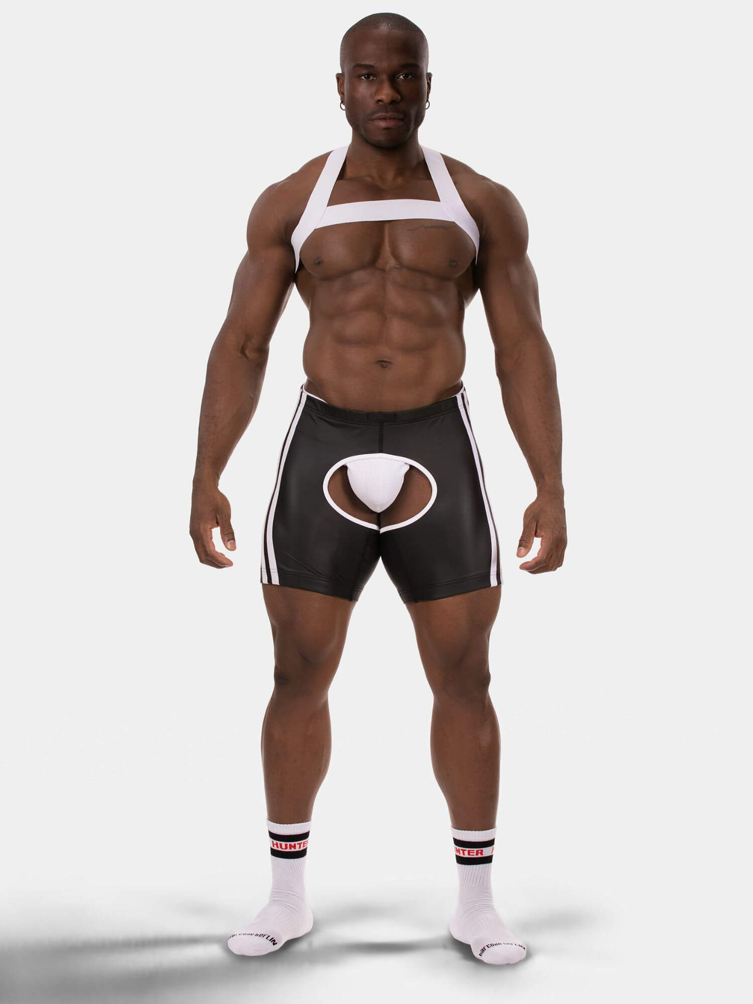 A muscular male model stands against a light gray studio background wearing a white halter-style harness top and black athletic shorts with white side stripes and a white circular front pouch. He is also wearing white socks.