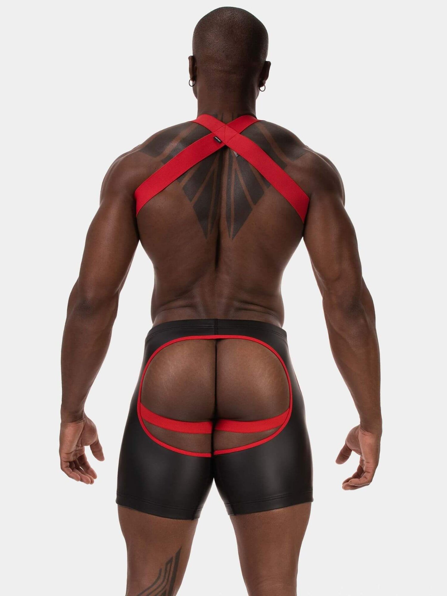 Rear view of a muscular man wearing black men's briefs with red elastic straps and a large rear cutout, set against a solid light gray background.