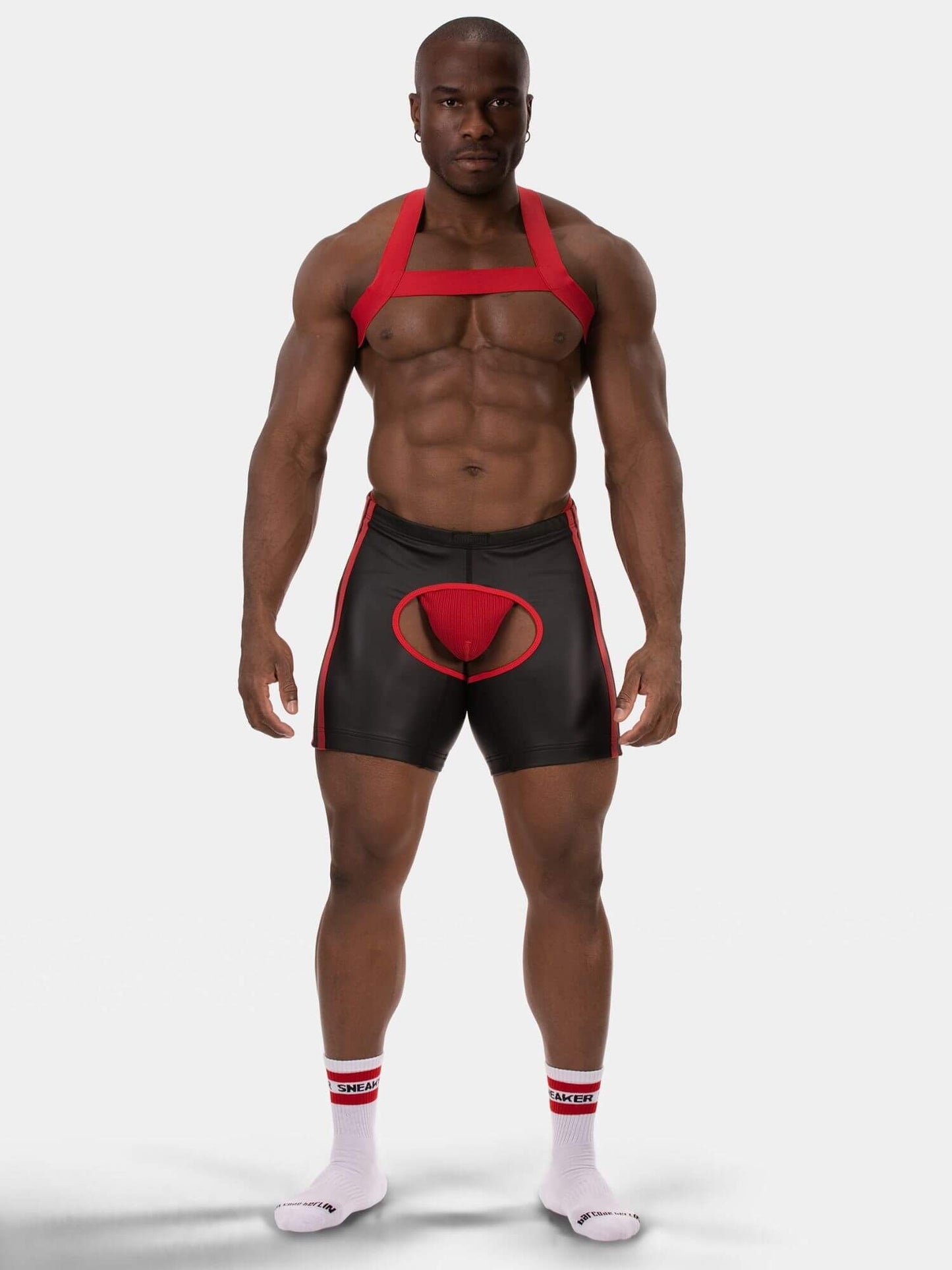 A muscular male model wearing black and red athletic briefs with a red pouch, a red harness-style top, and white socks with red stripes, photographed against a light gray studio background.