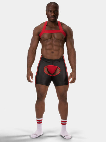 A muscular male model wearing black and red athletic briefs with a red pouch, a red harness-style top, and white socks with red stripes, photographed against a light gray studio background.