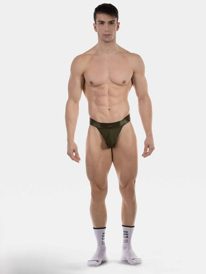 A muscular male model stands against a light gray studio background wearing a very minimal dark olive green thong-style brief and light gray socks with black text.