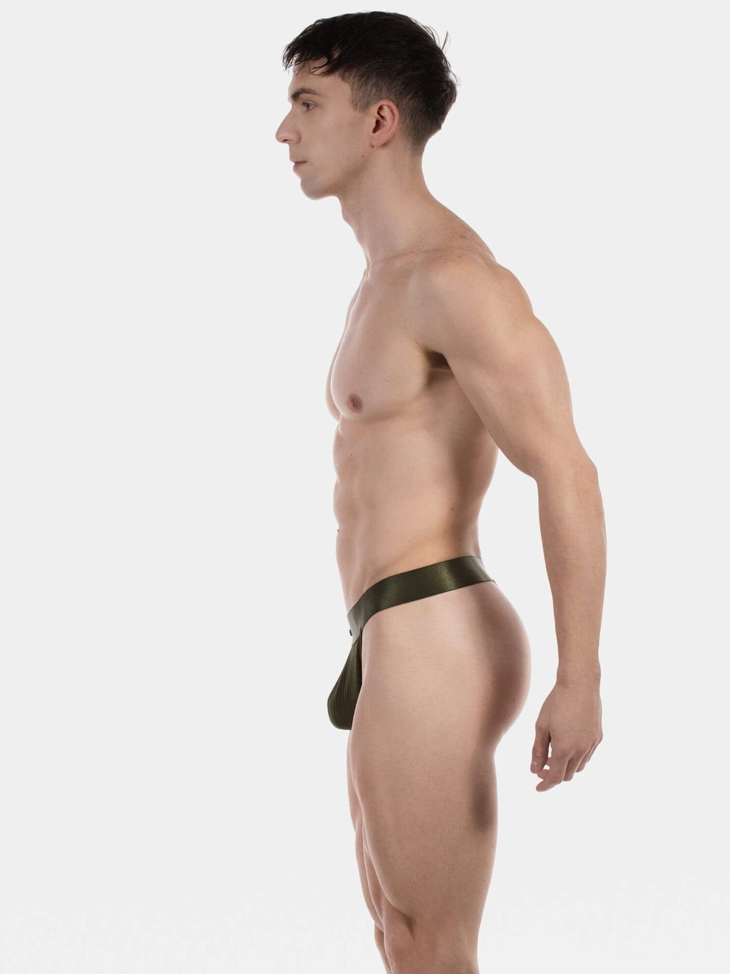 A muscular man in profile wearing a dark olive green minimal thong photographed against a seamless light gray studio background.