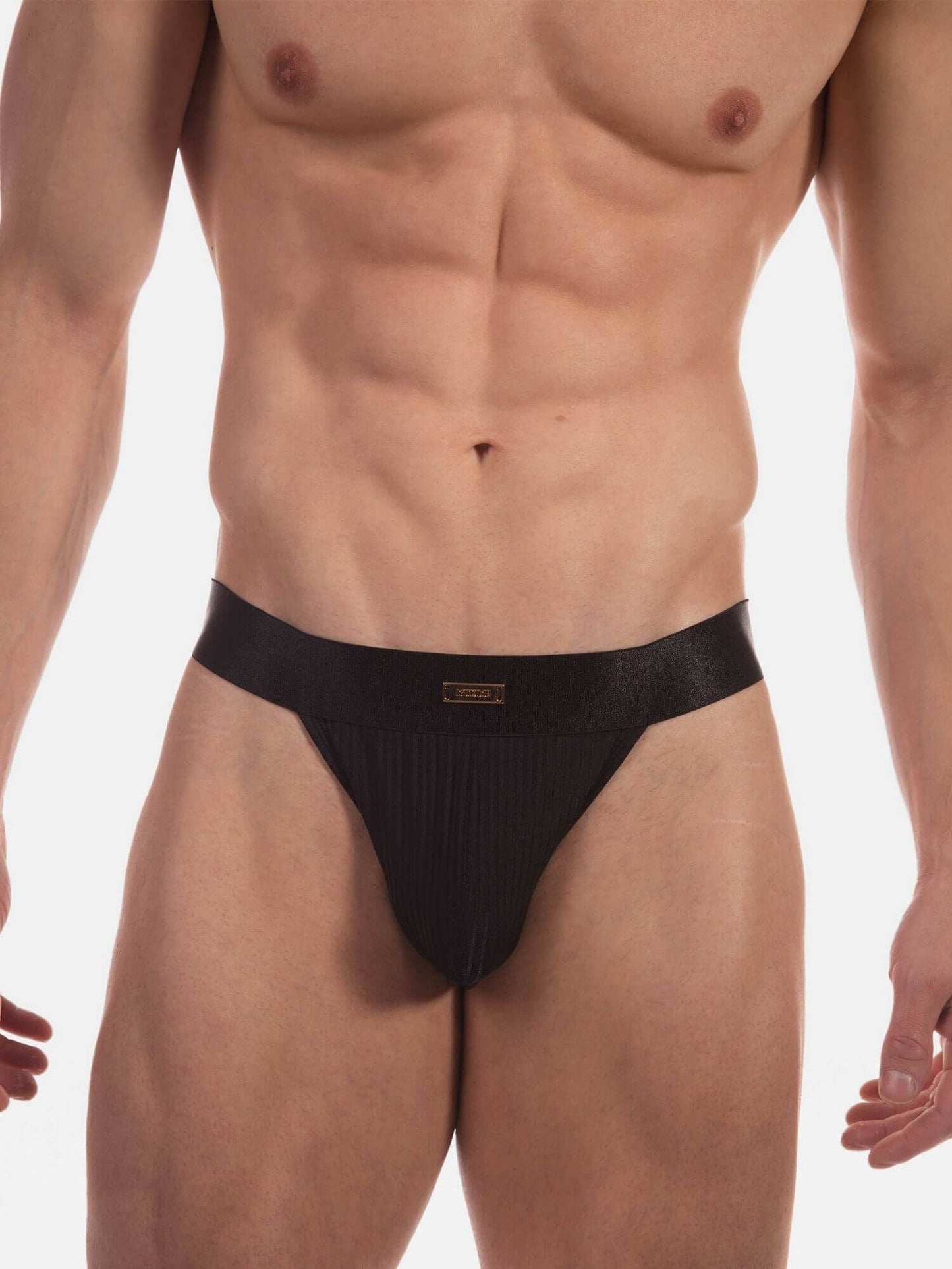 A close-up of a muscular male model wearing black ribbed men's thong against a plain white studio background.