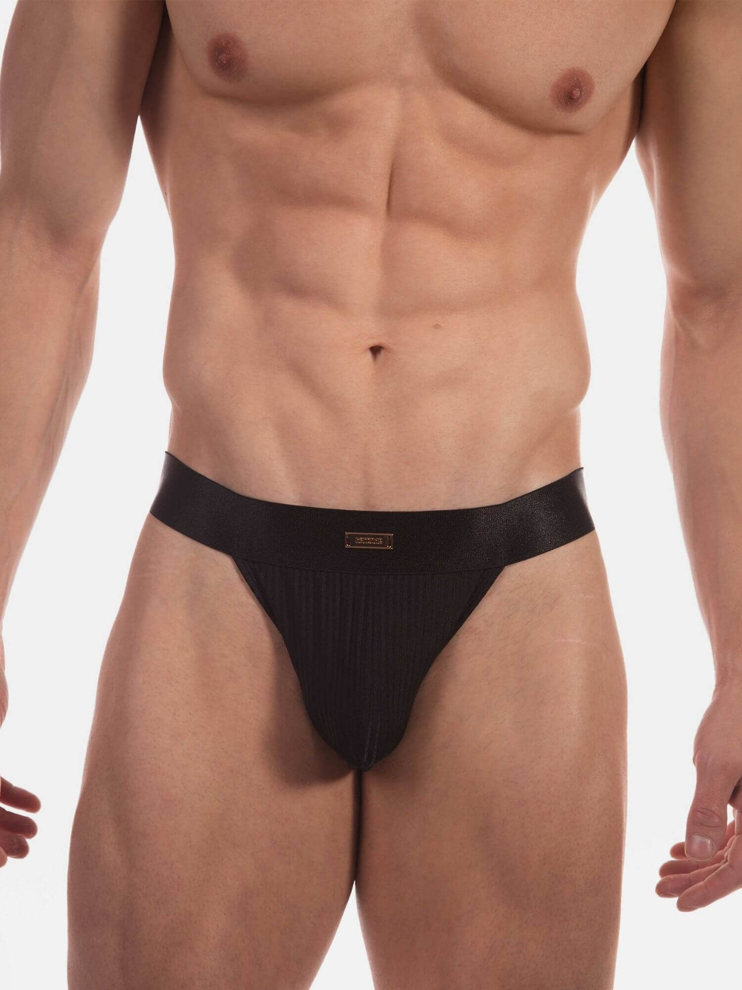 A close-up of a muscular male model wearing black ribbed men's thong against a plain white studio background.