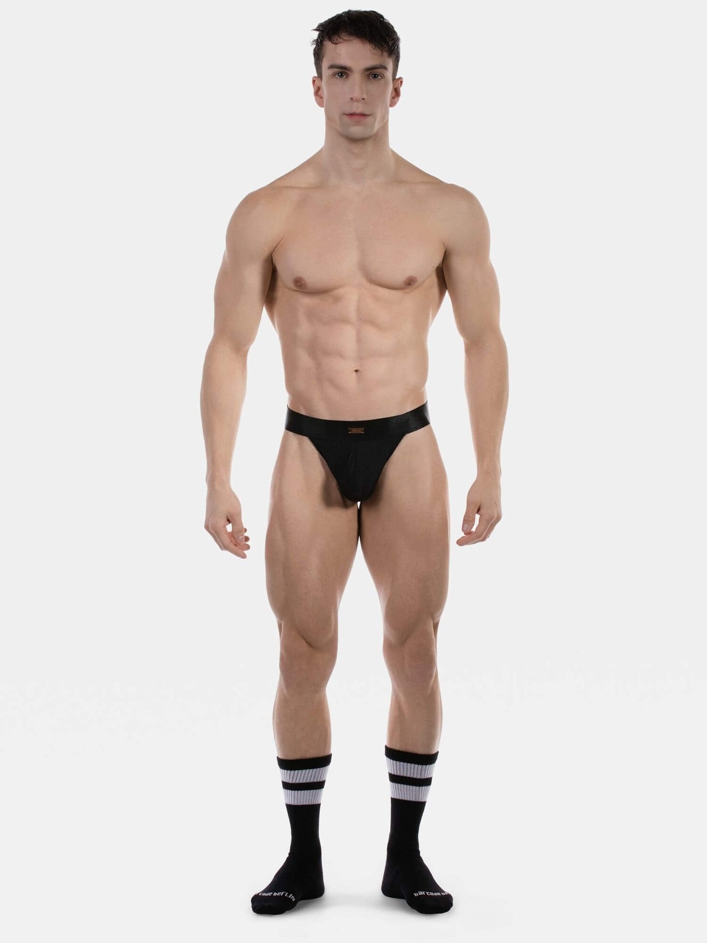 A muscular male model stands against a light gray studio background wearing a very minimal black thong  and black knee-high socks with white stripes.