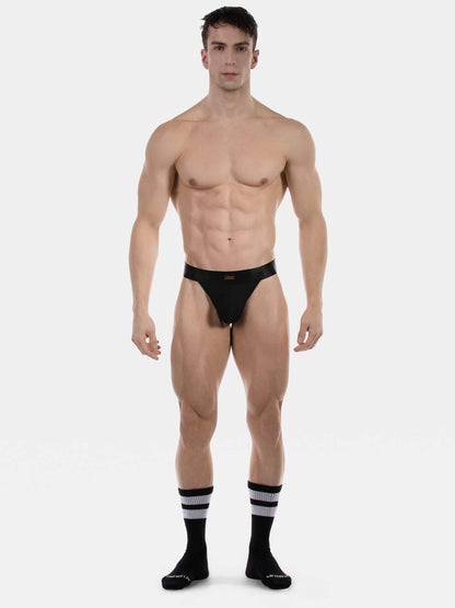 A muscular male model stands against a light gray studio background wearing a very minimal black thong  and black knee-high socks with white stripes.