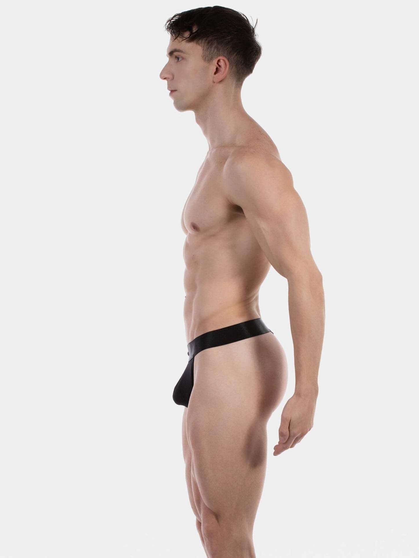 A side-profile view of a muscular man wearing a black men's thong photographed against a solid light gray studio background.