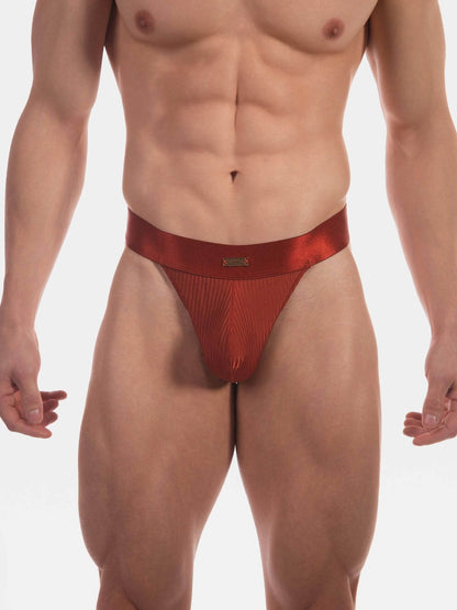 A close-up of a male model's lower torso wearing a metallic red thong with a textured front panel and a small gold brand tag at the center waistband, against a plain light gray studio background.