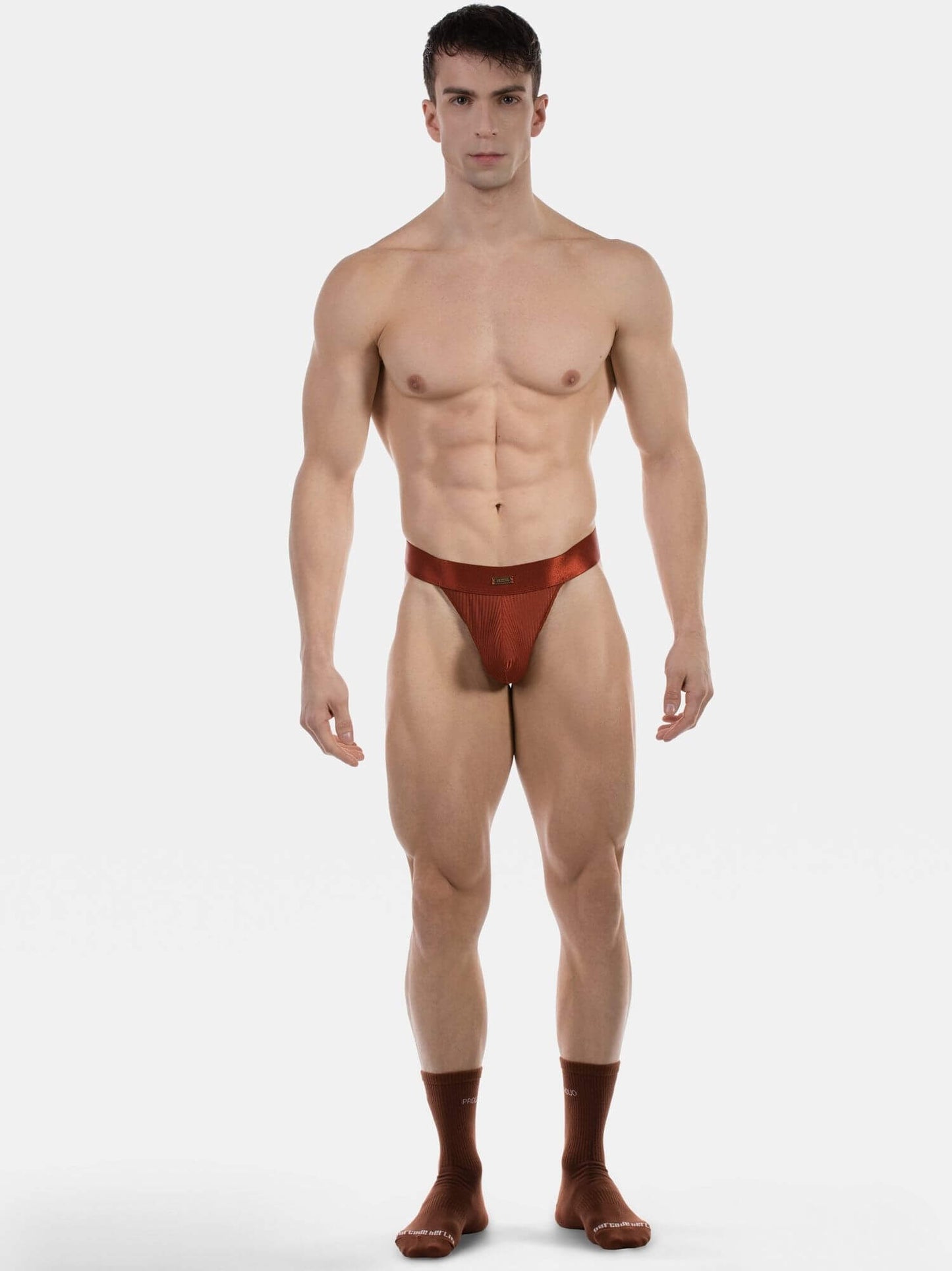 A muscular male model standing against a light gray studio background, wearing a very small red-brown thong-style brief and matching socks.