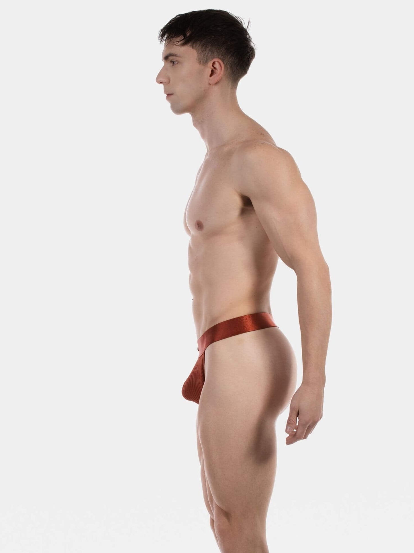 A man in profile wearing a satin red men's thong, photographed against a solid light gray background.