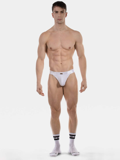 A muscular male model stands against a light gray studio background wearing a very minimal white thong and white socks with black stripes.