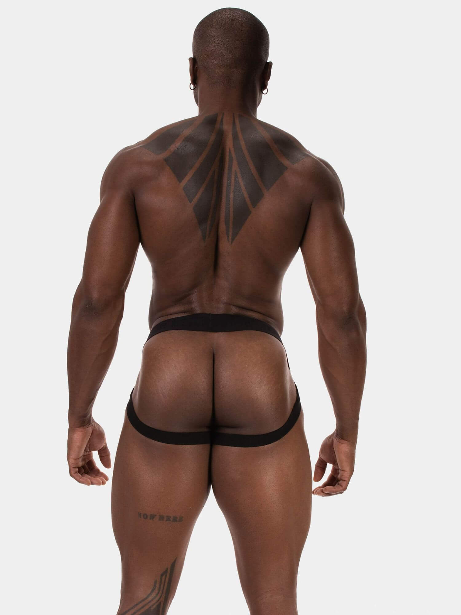 Rear view of a muscular man with tattoos wearing black men's briefs against a light gray studio background.