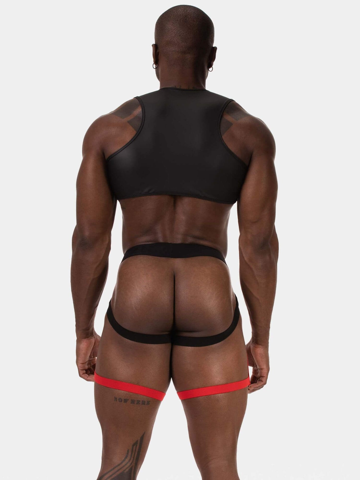 Rear view of a muscular man wearing black athletic underwear and a red garter belt against a light gray studio background.