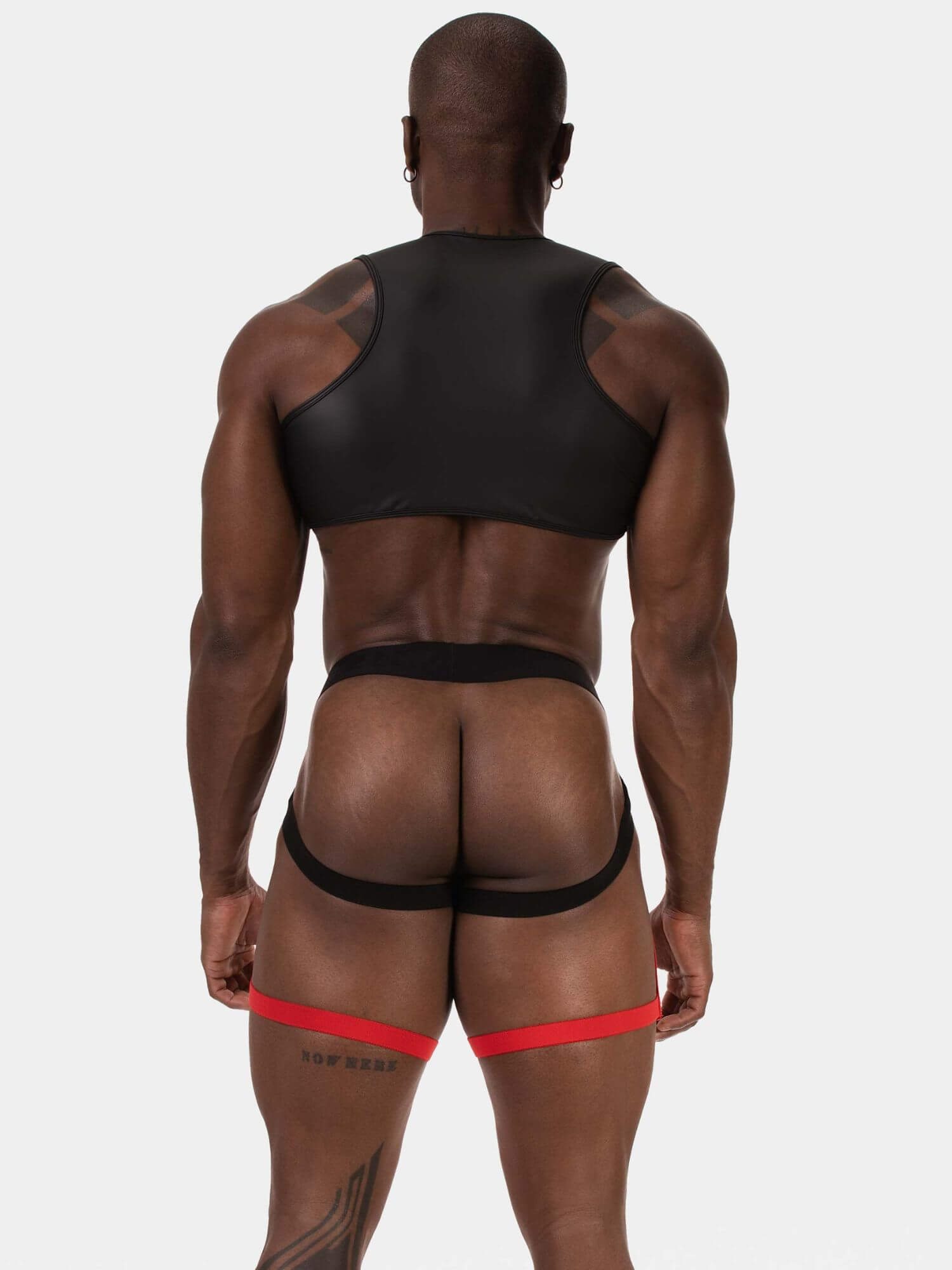 Rear view of a muscular man wearing black athletic underwear and a red garter belt against a light gray studio background.