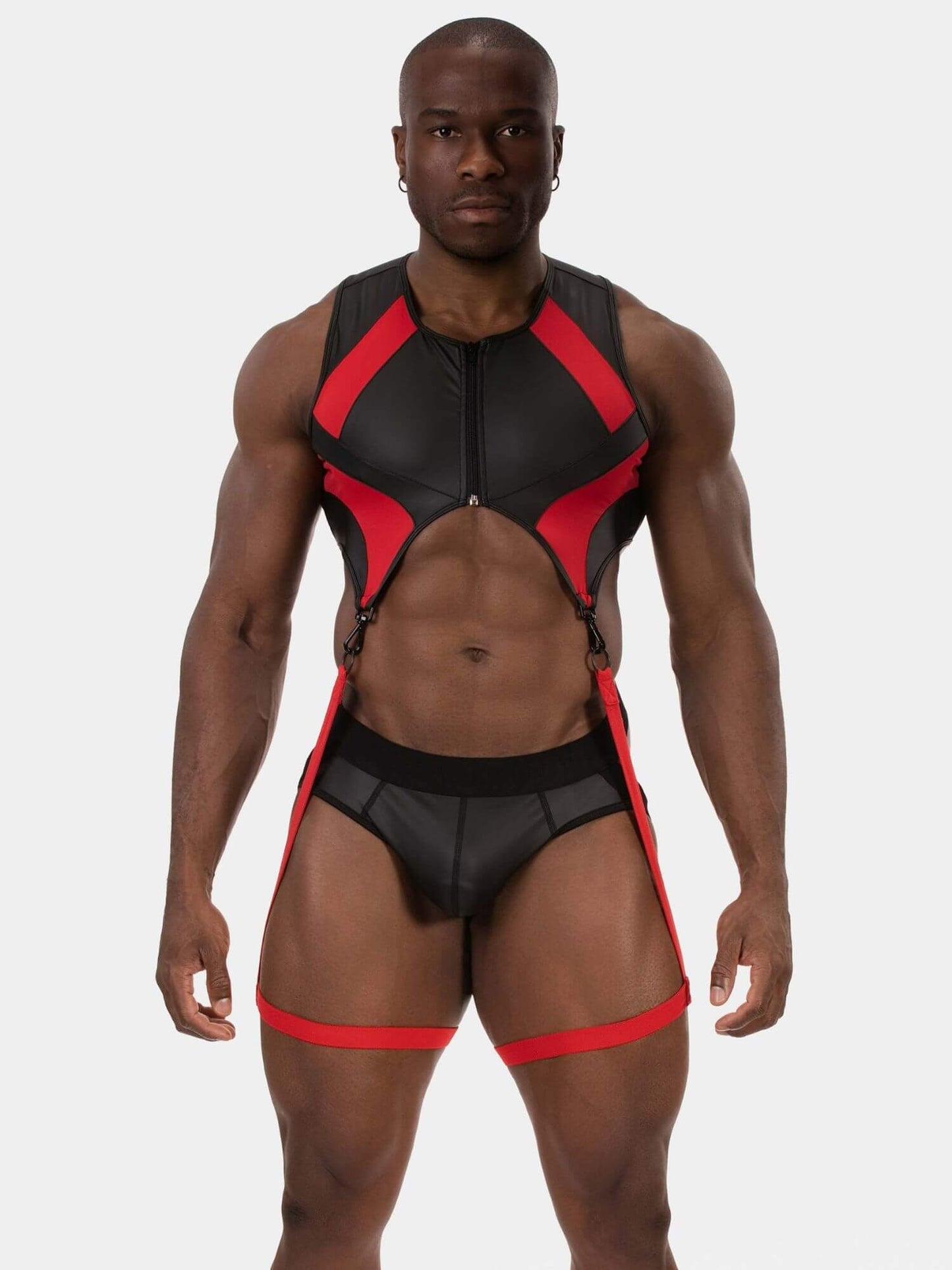A muscular male model wearing a black and red strappy bodysuit with a front zipper and matching briefs, photographed against a light gray studio background.