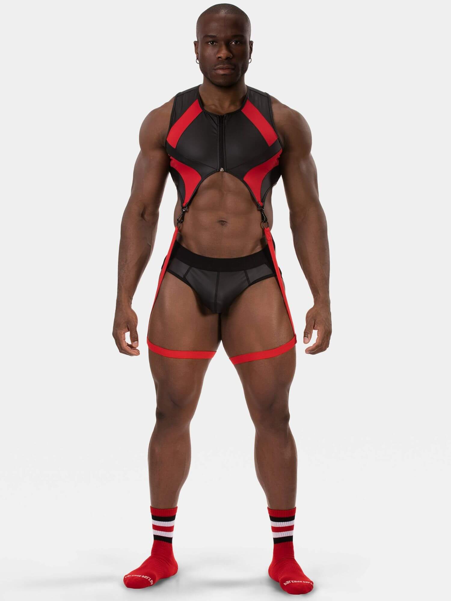 A muscular male model wearing a black and red athletic harness-style bodysuit with matching briefs and red knee-high socks, photographed against a light gray studio background.