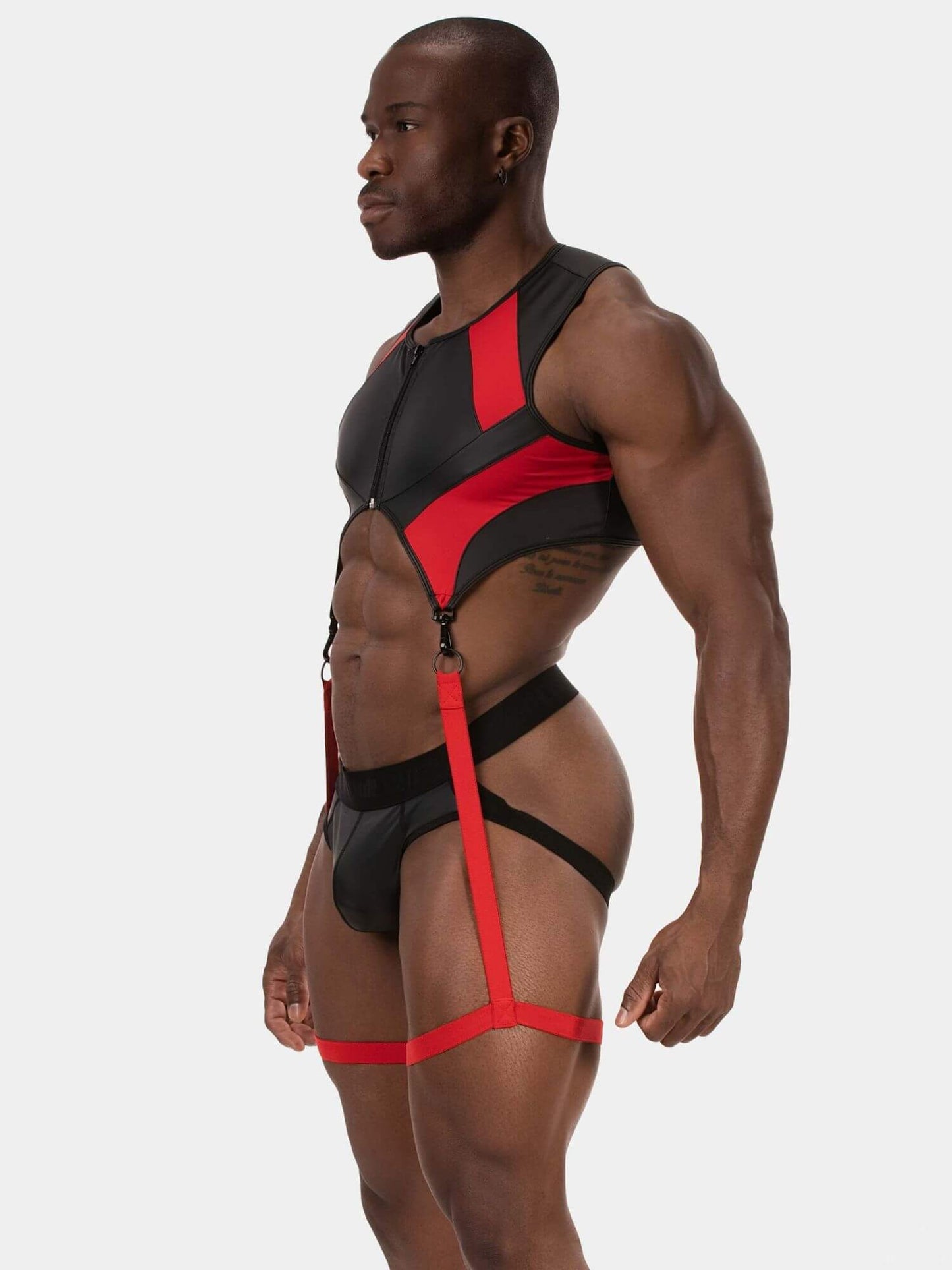 A muscular man wearing a black and red strappy harness-style brief set, shown in profile against a light gray studio background.