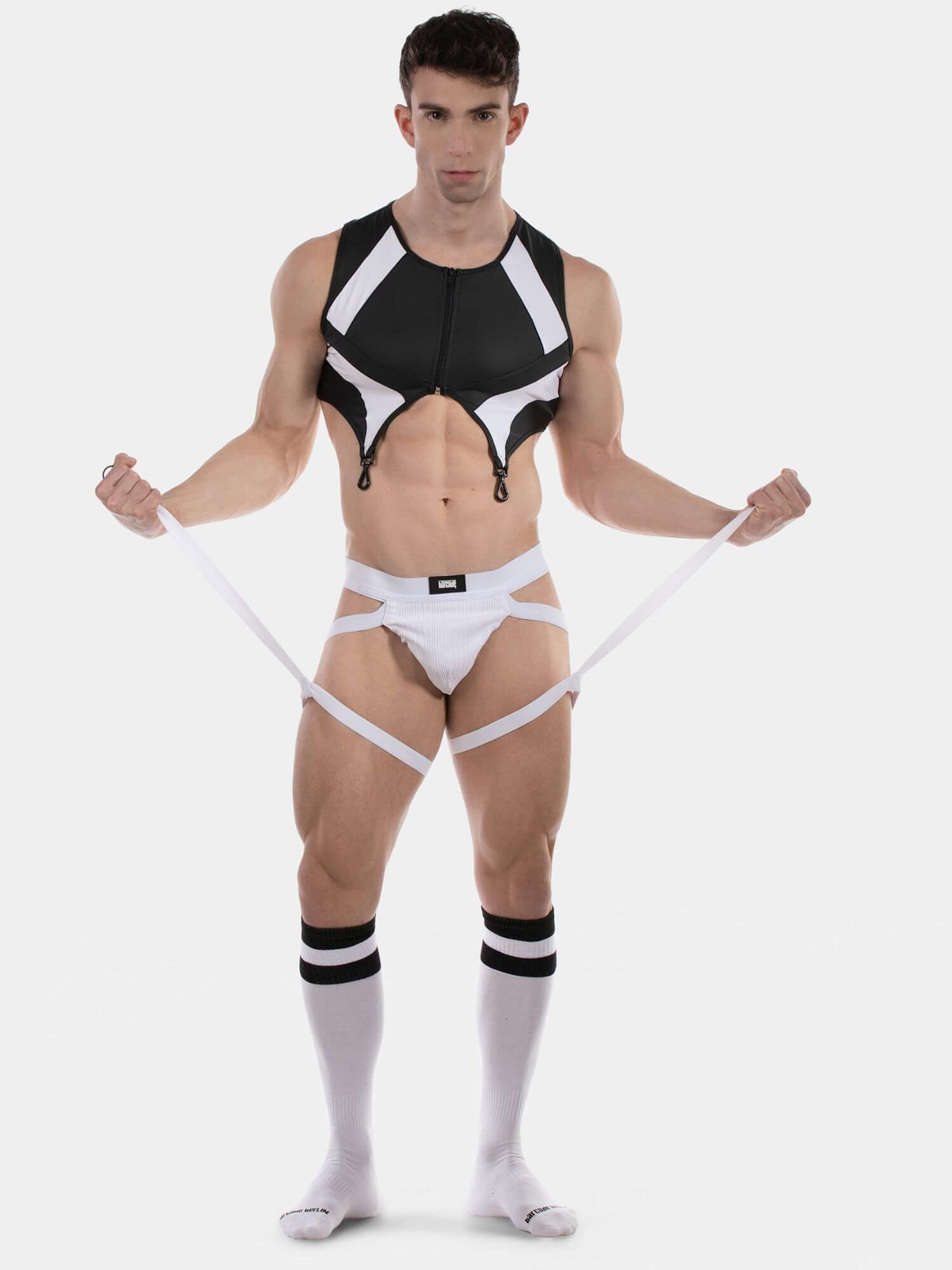 A muscular man modeling a black and white strappy men's harness-style lingerie set, including a cropped vest, white briefs, and white knee-high socks with black stripes, against a light gray background.