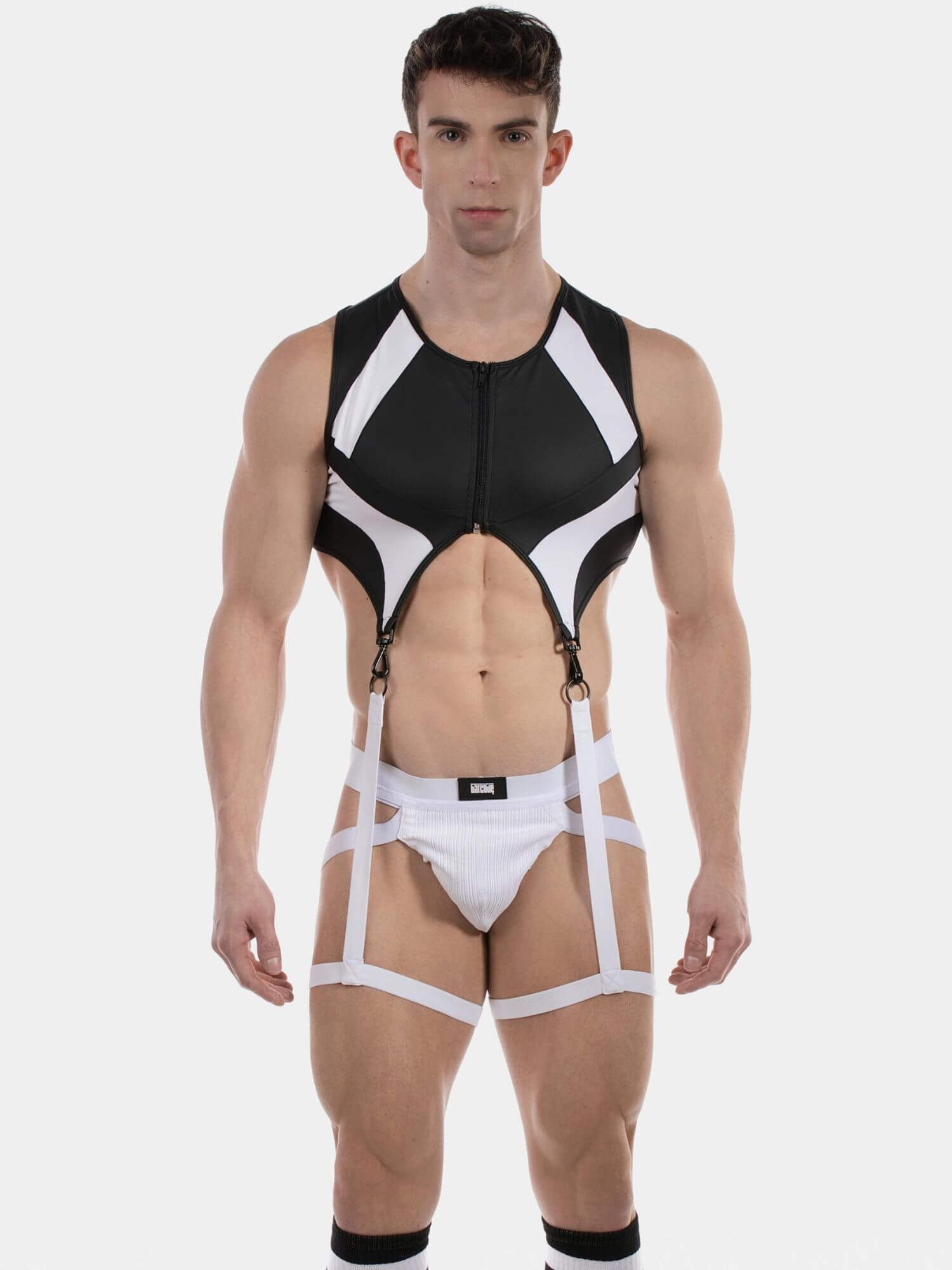 A muscular male model wearing a black and white strappy harness-style bodysuit with a front zipper, white briefs, and black knee socks, photographed against a light gray studio background.