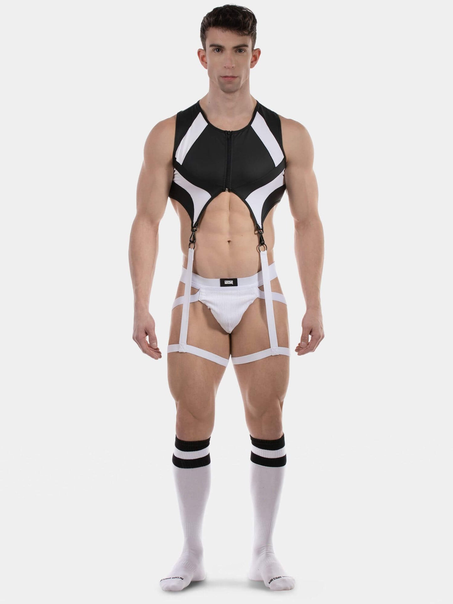A muscular male model wearing a black and white strappy harness-style bodysuit with integrated briefs, suspenders, and matching white knee-high socks with black stripes, photographed against a light gray studio background.