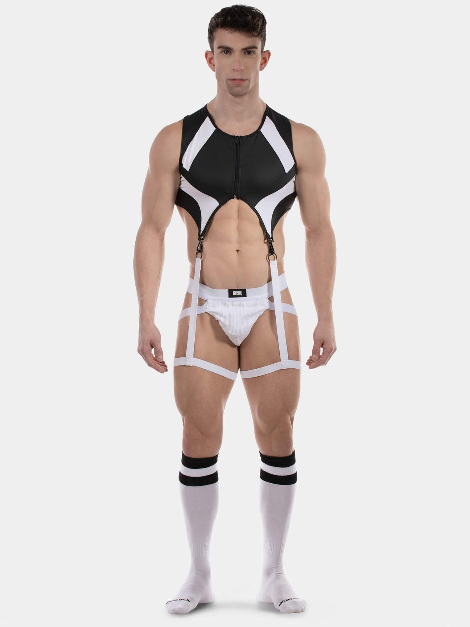 A muscular male model wearing a black and white strappy harness-style bodysuit with integrated briefs, suspenders, and matching white knee-high socks with black stripes, photographed against a light gray studio background.
