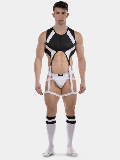 A muscular male model wearing a black and white strappy harness-style bodysuit with integrated briefs, suspenders, and matching white knee-high socks with black stripes, photographed against a light gray studio background.