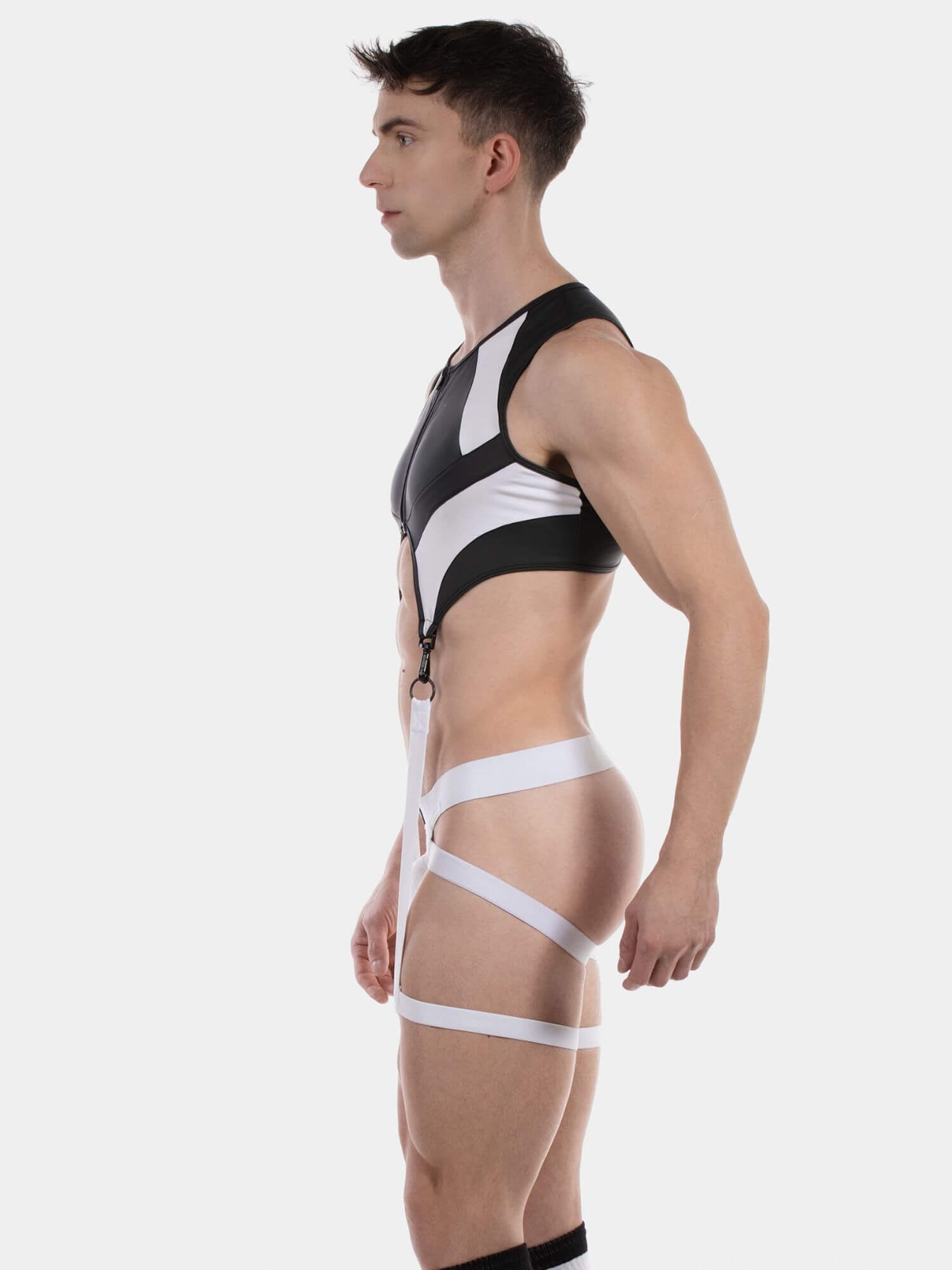 A man in side profile wearing a black and white strappy athletic support garment with a cropped tank top and thigh straps, photographed against a light gray studio background.