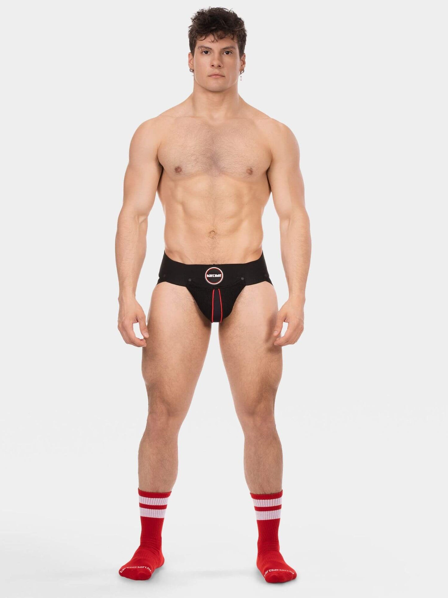A shirtless male model stands against a light gray studio background wearing black jockstrap with a red center stripe and a circular logo at the front, along with red socks with white stripes.