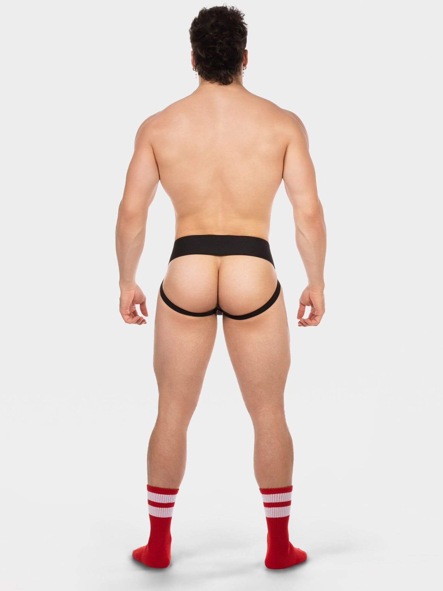 A muscular man is shown from behind wearing black men's briefs and red crew socks with white stripes, standing against a light gray studio background.