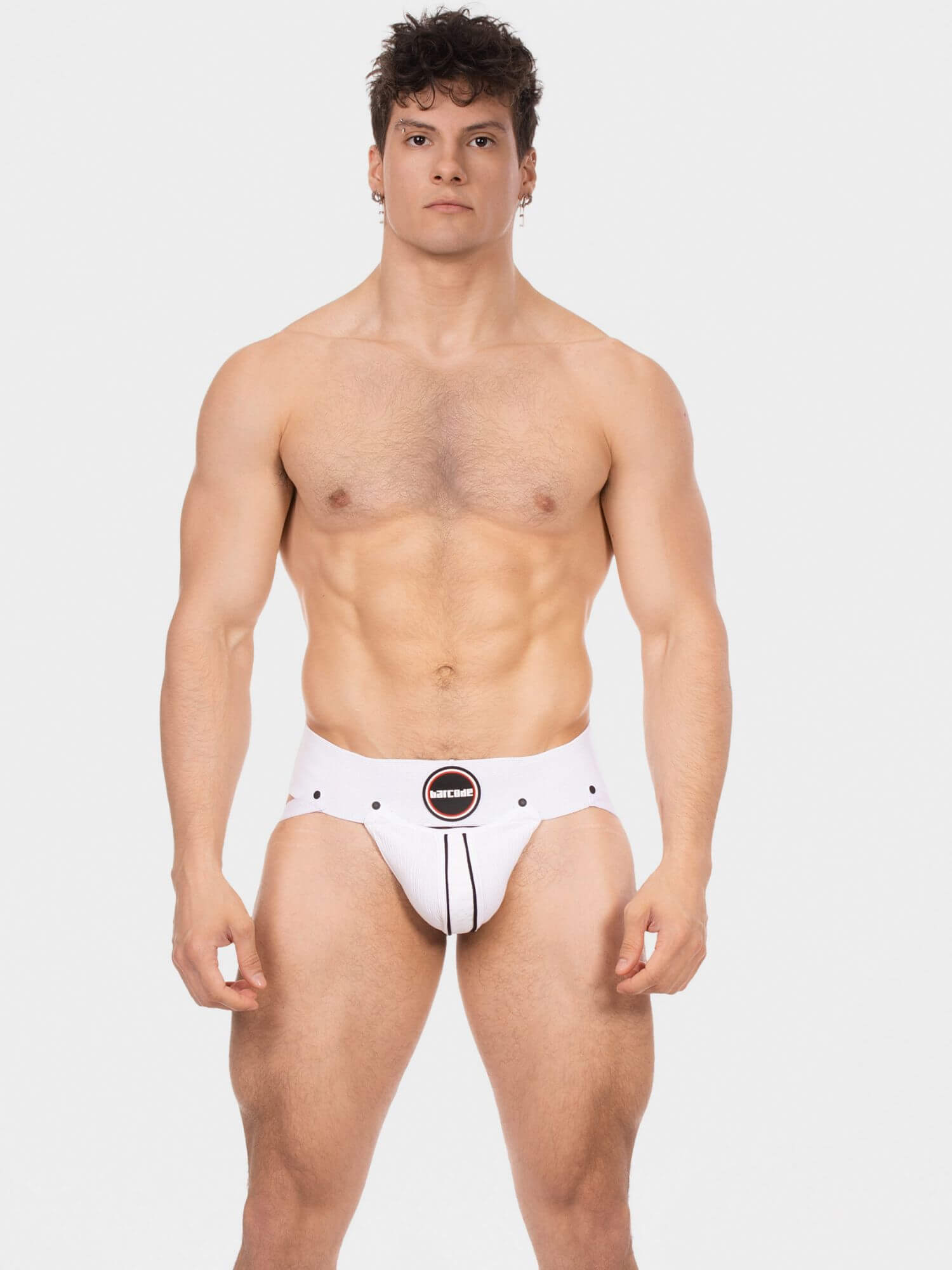 A muscular man modeling a white men's jockstrap with black trim and a circular black-and-white logo patch at the center front, photographed against a light gray studio background.