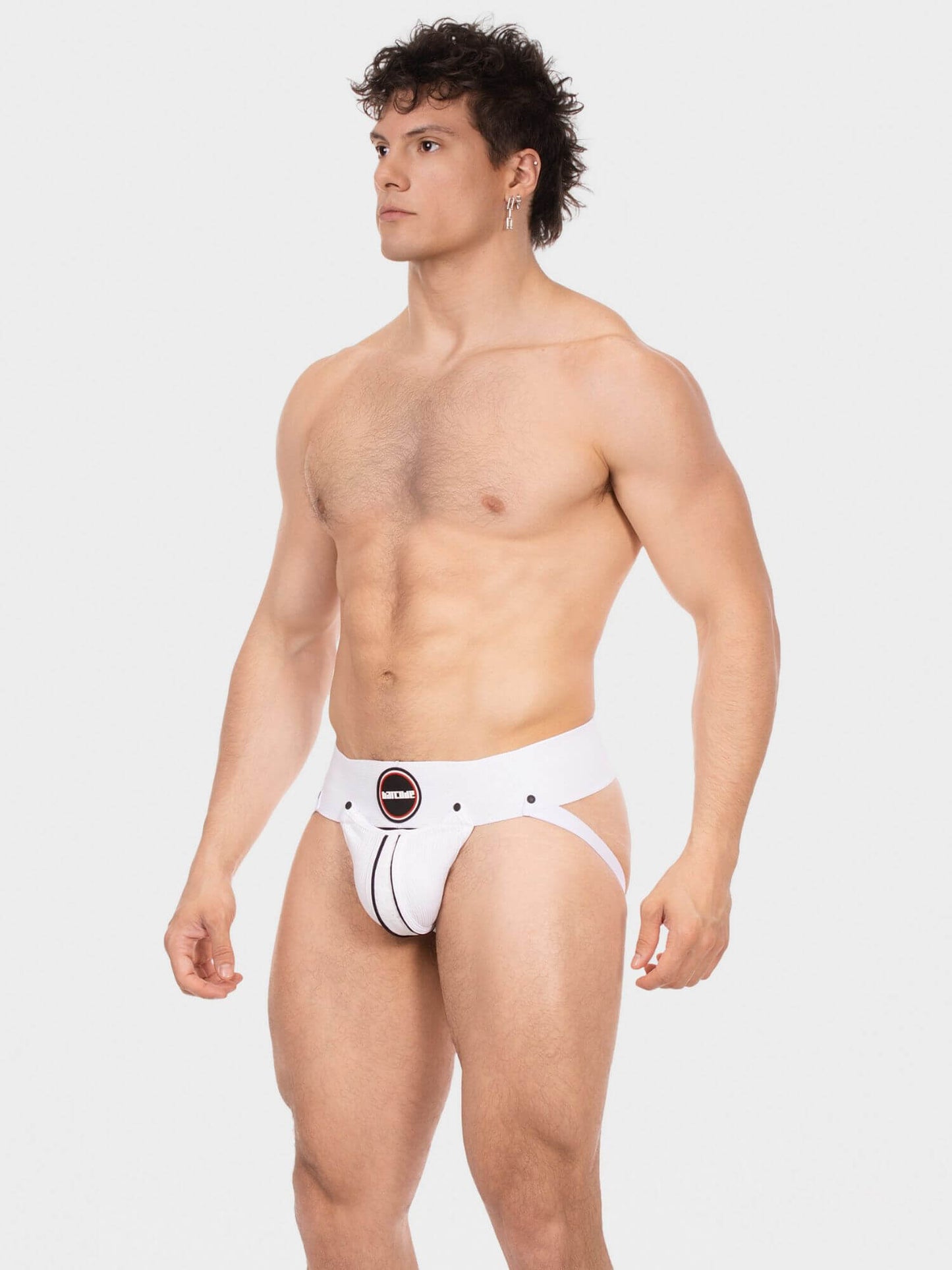 A shirtless male model wearing white men's briefs with a black circular logo patch on the front, photographed against a light gray studio background.