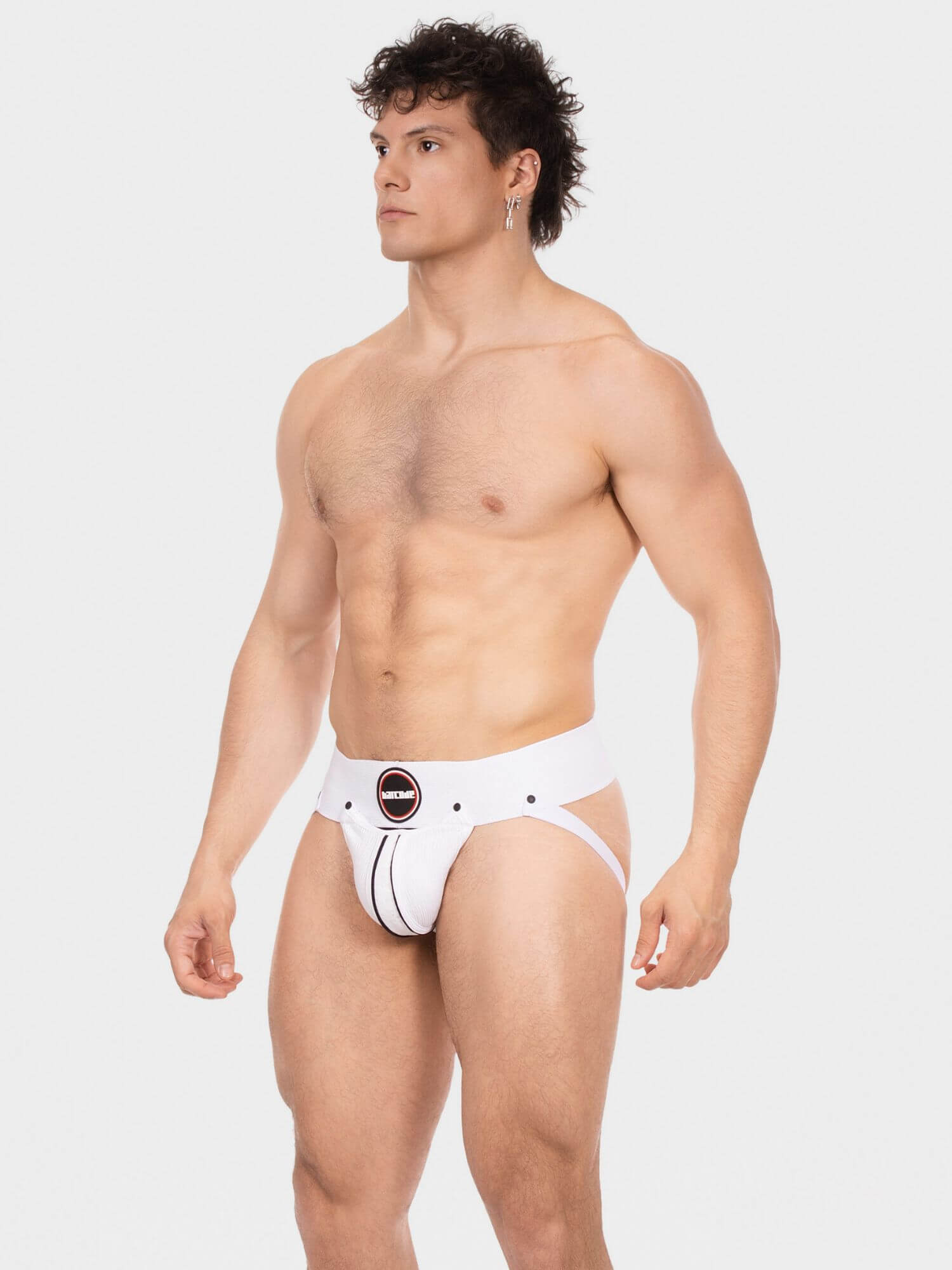 A shirtless male model wearing white men's briefs with a black circular logo patch on the front, photographed against a light gray studio background.