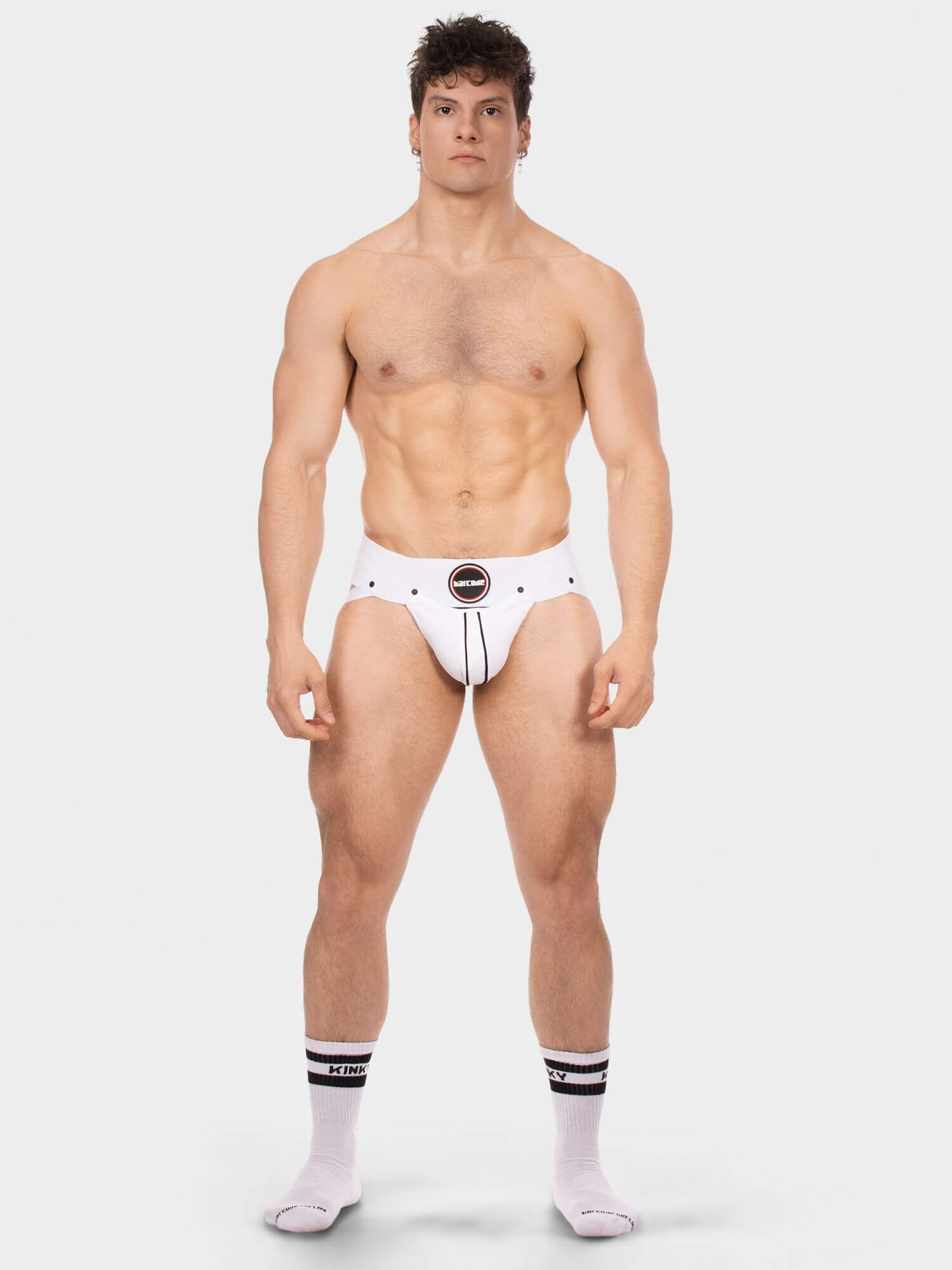 A muscular male model stands against a light gray studio background wearing white briefs with a black circular logo at the center front and white crew socks with black stripes.